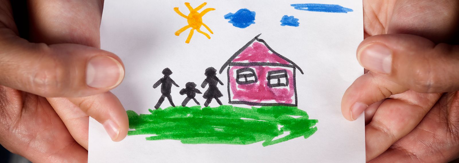 Hände halten ein Papier auf dem eine Kinderzeichnung mit einem Haus und drei Personen zu sehen ist