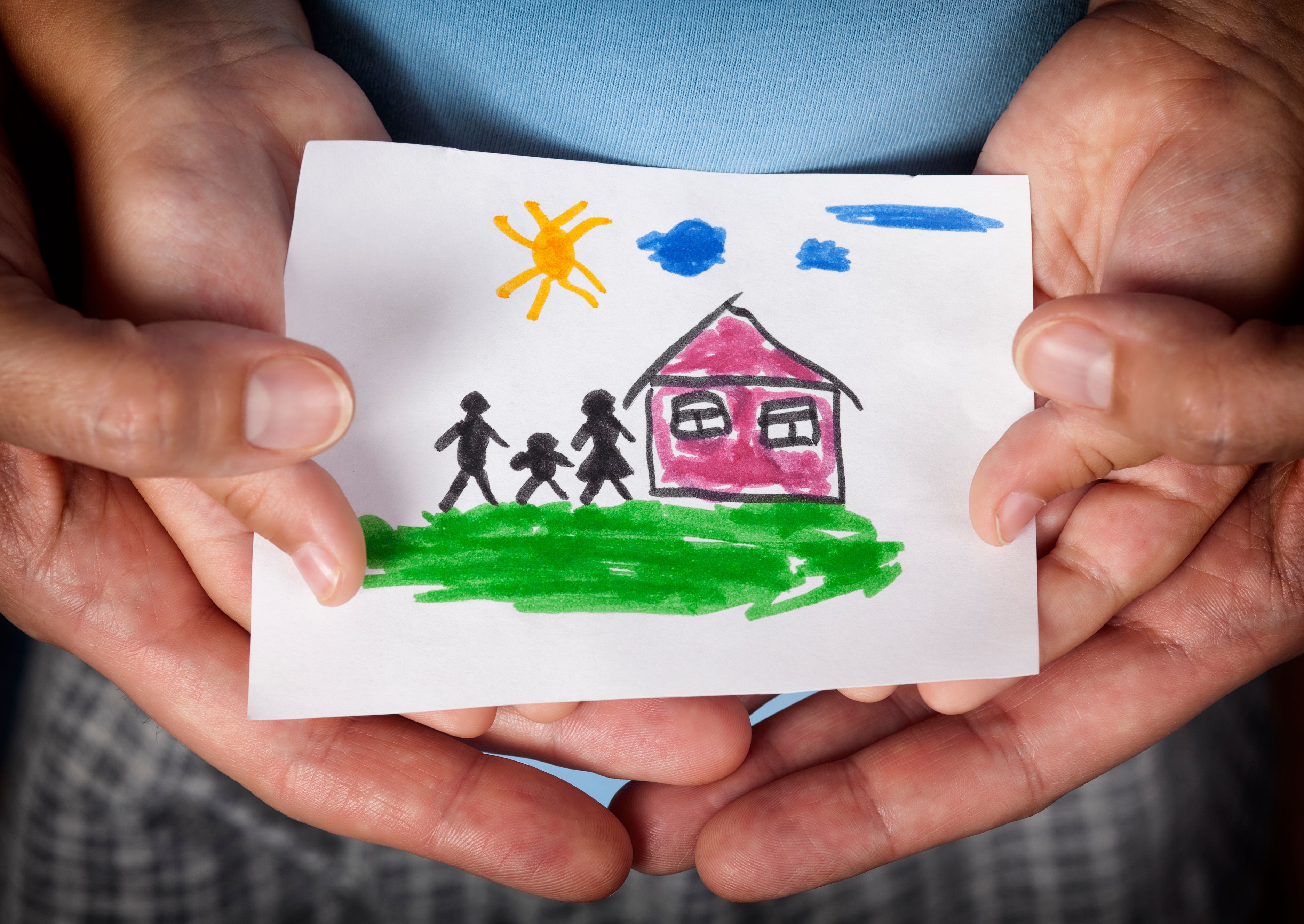 Hände halten ein Papier auf dem eine Kinderzeichnung mit einem Haus und drei Personen zu sehen ist